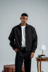 Man wearing a black jacket and pants standing in a room with a table and lamp.