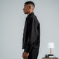 Man in a black outfit standing next to a suitcase with a small table and lamp in the background.