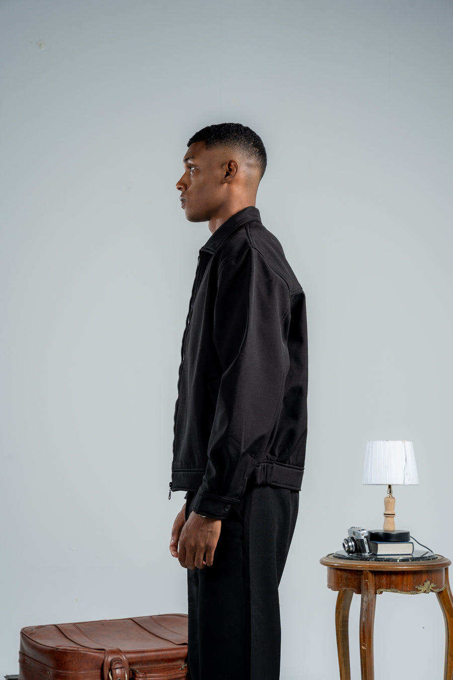 Man in a black outfit standing next to a suitcase with a small table and lamp in the background.