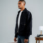 Man wearing a black jacket and blue jeans standing next to vintage suitcases against a plain background