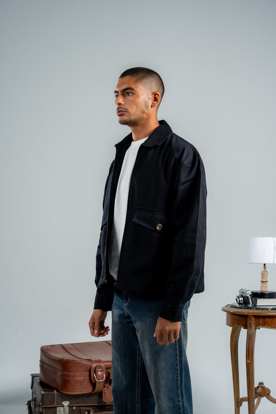 Man wearing a black jacket and blue jeans standing next to vintage suitcases against a plain background