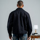 Man wearing a black jacket and blue jeans standing in front of a gray wall with a small table and lamp in the background.