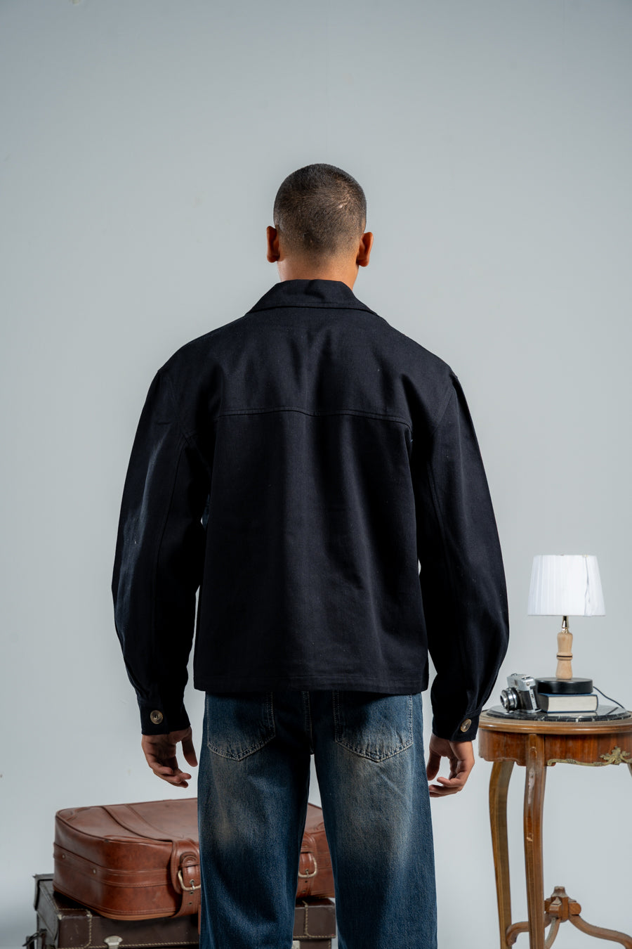 Man wearing a black jacket and blue jeans standing in front of a gray wall with a small table and lamp in the background.