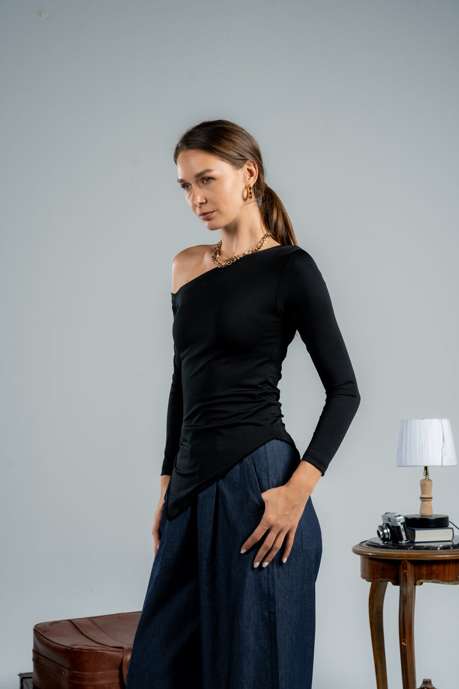 Woman wearing a black one-shoulder top and blue pants against a gray background