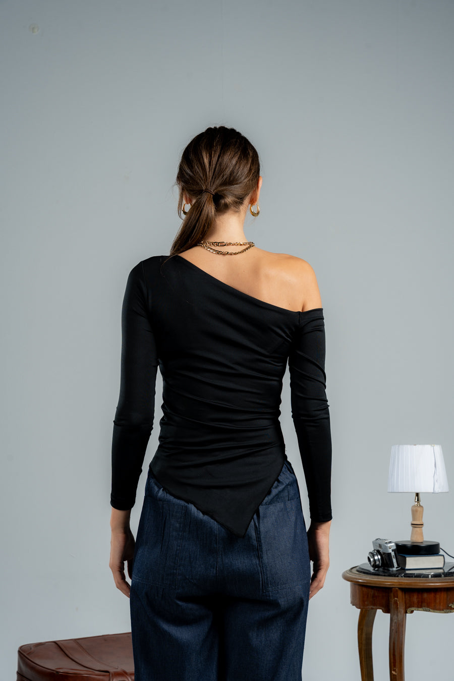 Woman wearing a black one-shoulder top and blue pants against a gray background