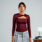 Woman wearing a burgundy long-sleeve top with cut-out design and light gray pants, standing against a plain background.