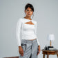 Woman wearing a white long-sleeve top with cut-out design and gray pants, standing next to a suitcase and small table.