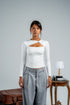 Woman wearing a white long-sleeve top with a cutout design and gray pants against a plain background.