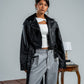 Person wearing a black leather jacket over a white top and gray pants, standing in a room with a suitcase and table.