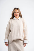 Woman wearing a beige sweater and pants with a white shirt underneath on a white background