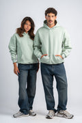 Two people wearing light green hoodies and dark jeans on a white background