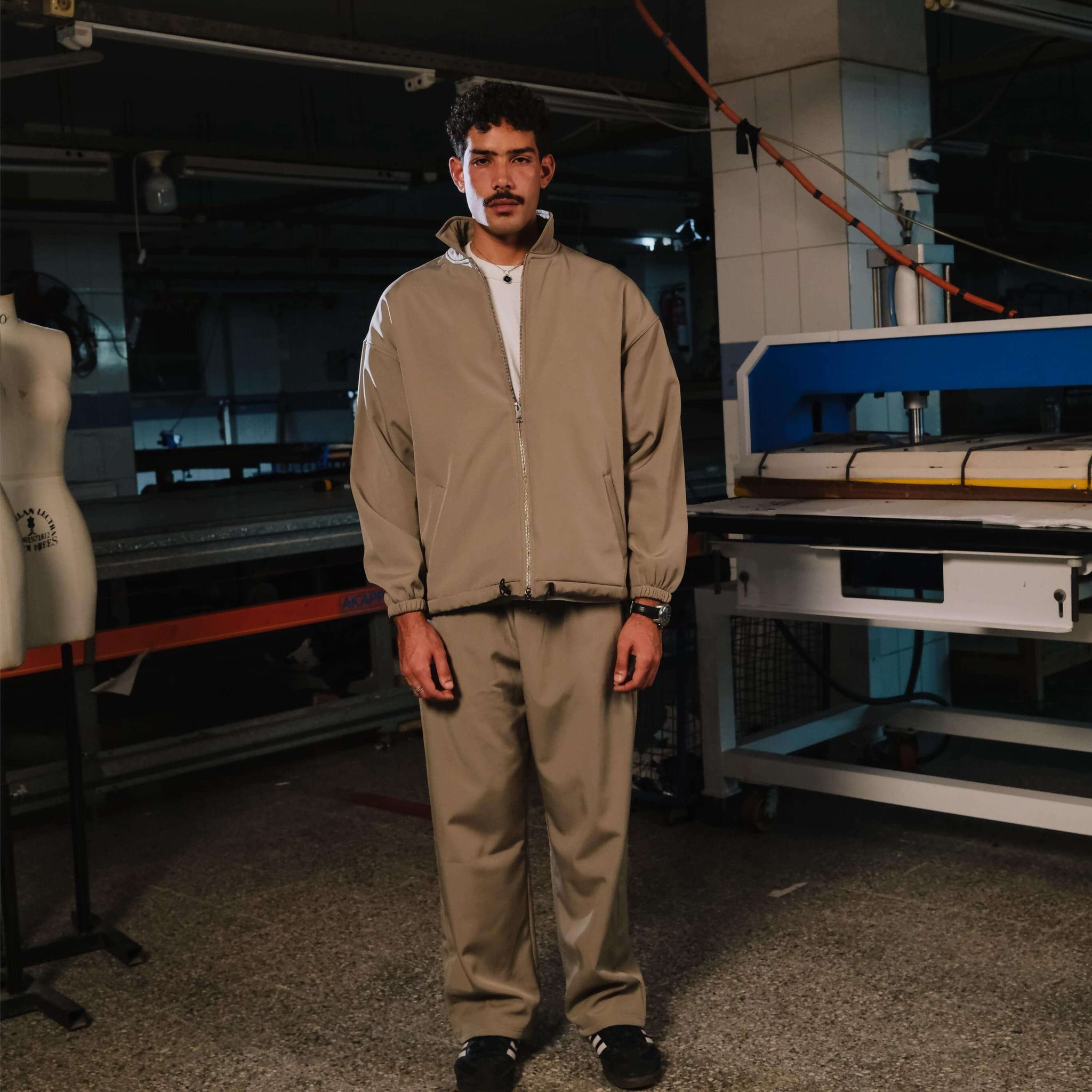 Person wearing a beige tracksuit standing in an industrial setting with machinery.