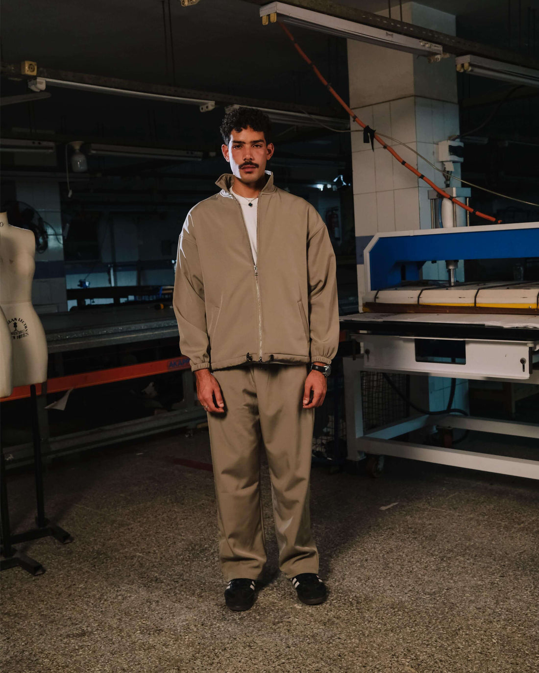 Person wearing a beige tracksuit standing in an industrial setting with machinery.