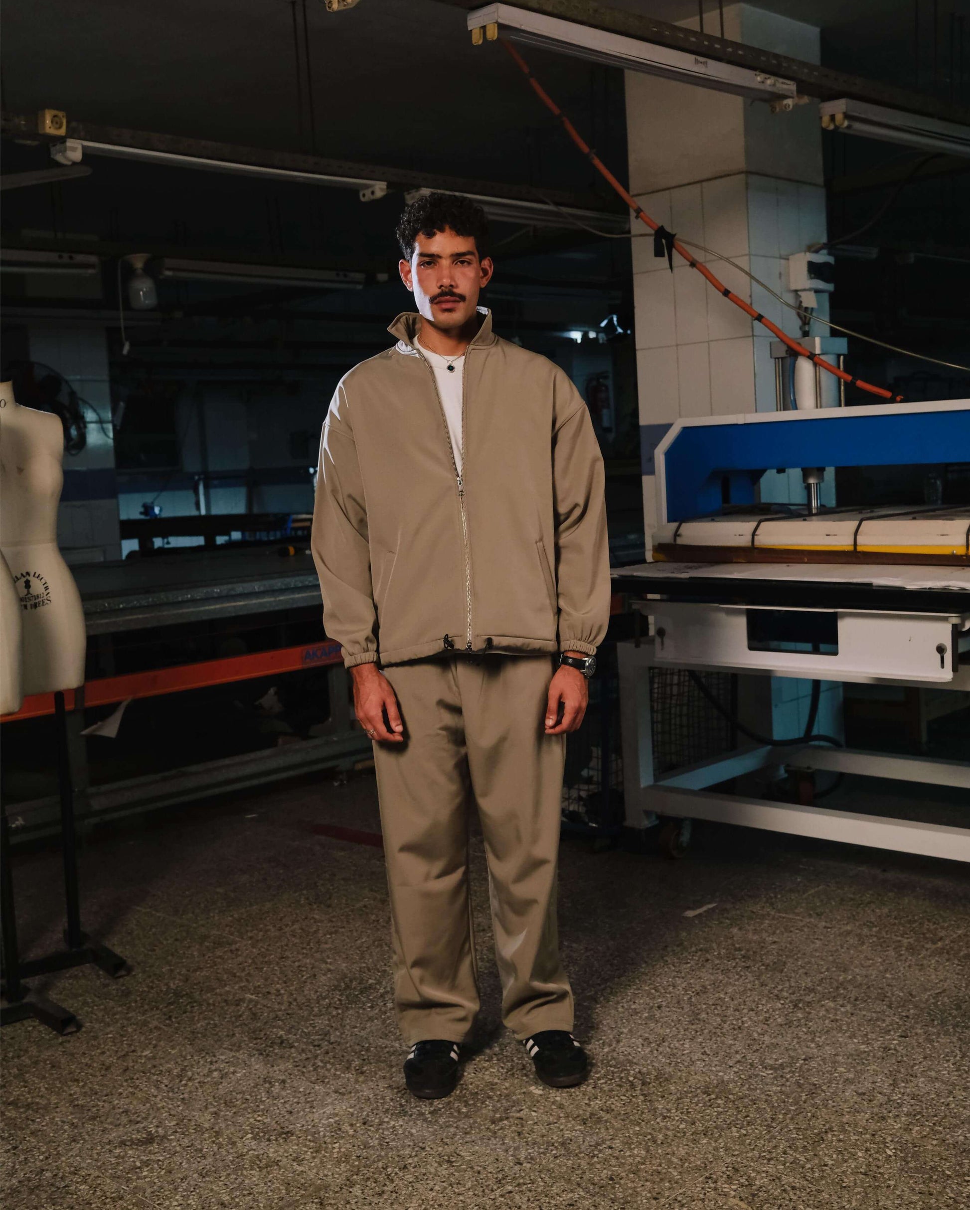 Person wearing a beige tracksuit standing in an industrial setting with machinery.