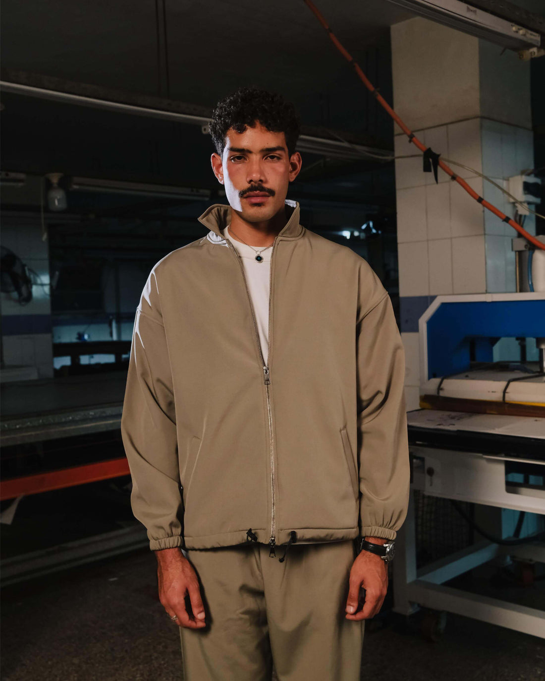 Person wearing a beige tracksuit standing in an industrial setting with machinery.