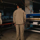 Person wearing a beige work uniform standing in an industrial setting with large equipment.