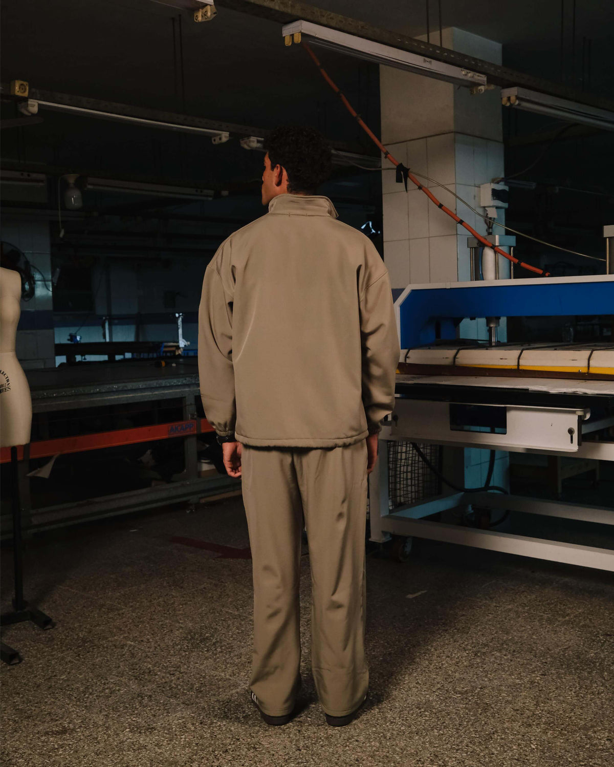 Person wearing a beige work uniform standing in an industrial setting with large equipment.