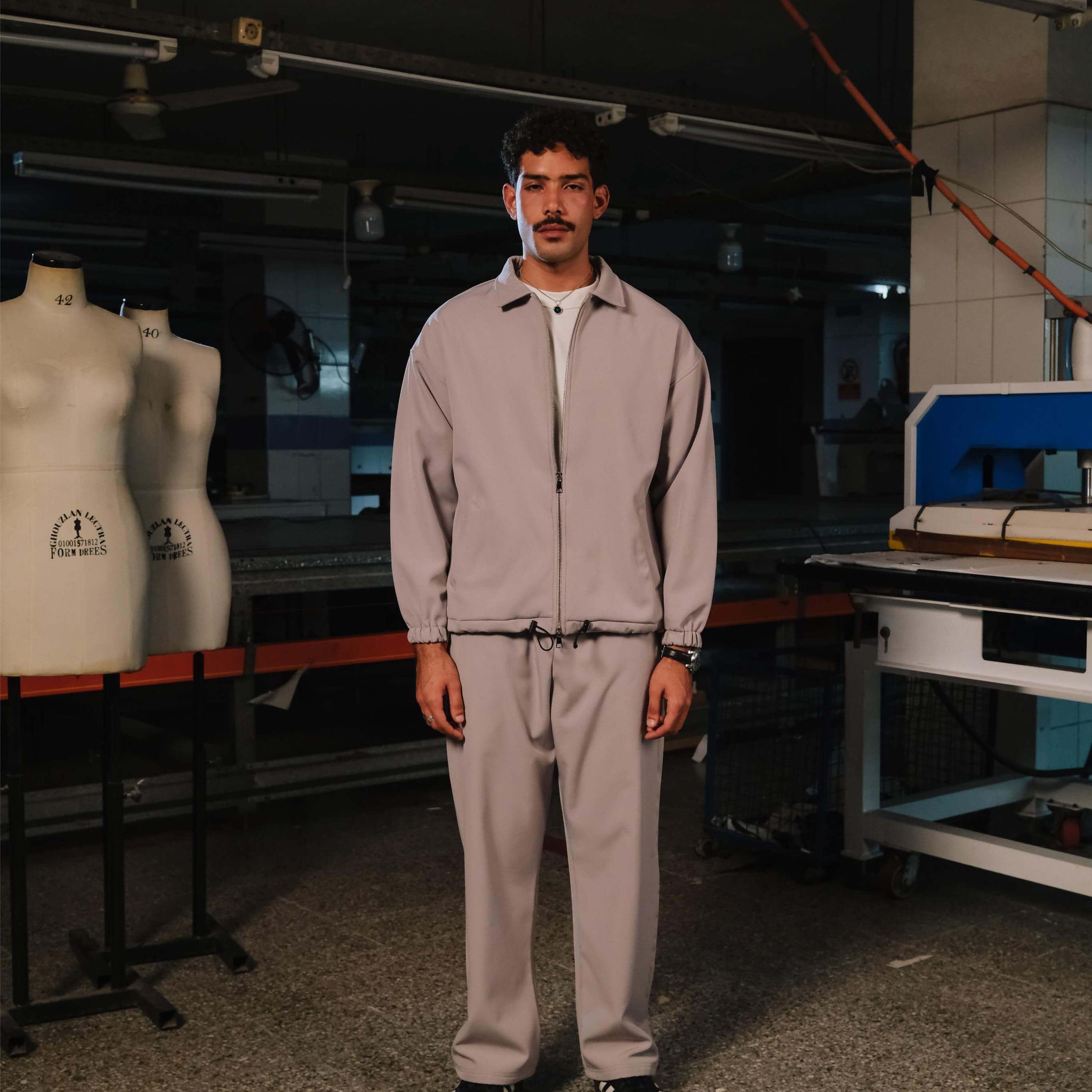 Person wearing a light gray tracksuit standing in a workshop with mannequins and equipment in the background.