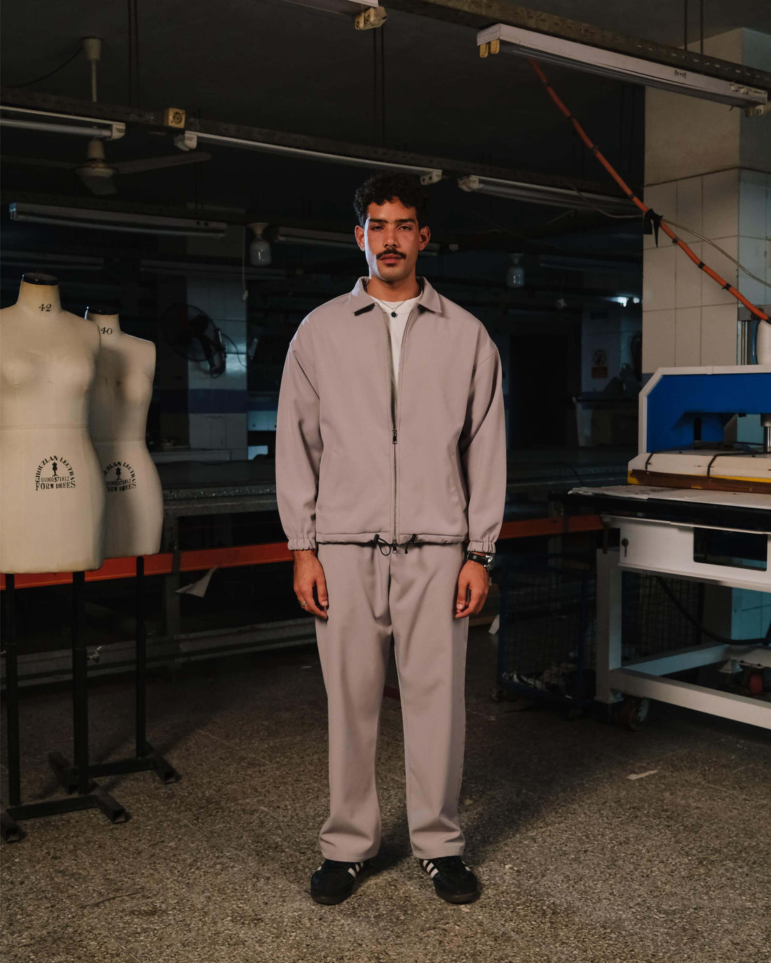Person wearing a light gray tracksuit standing in a workshop with mannequins and equipment in the background.