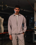 Man wearing a light gray tracksuit standing in a dark room with mannequin and equipment.
