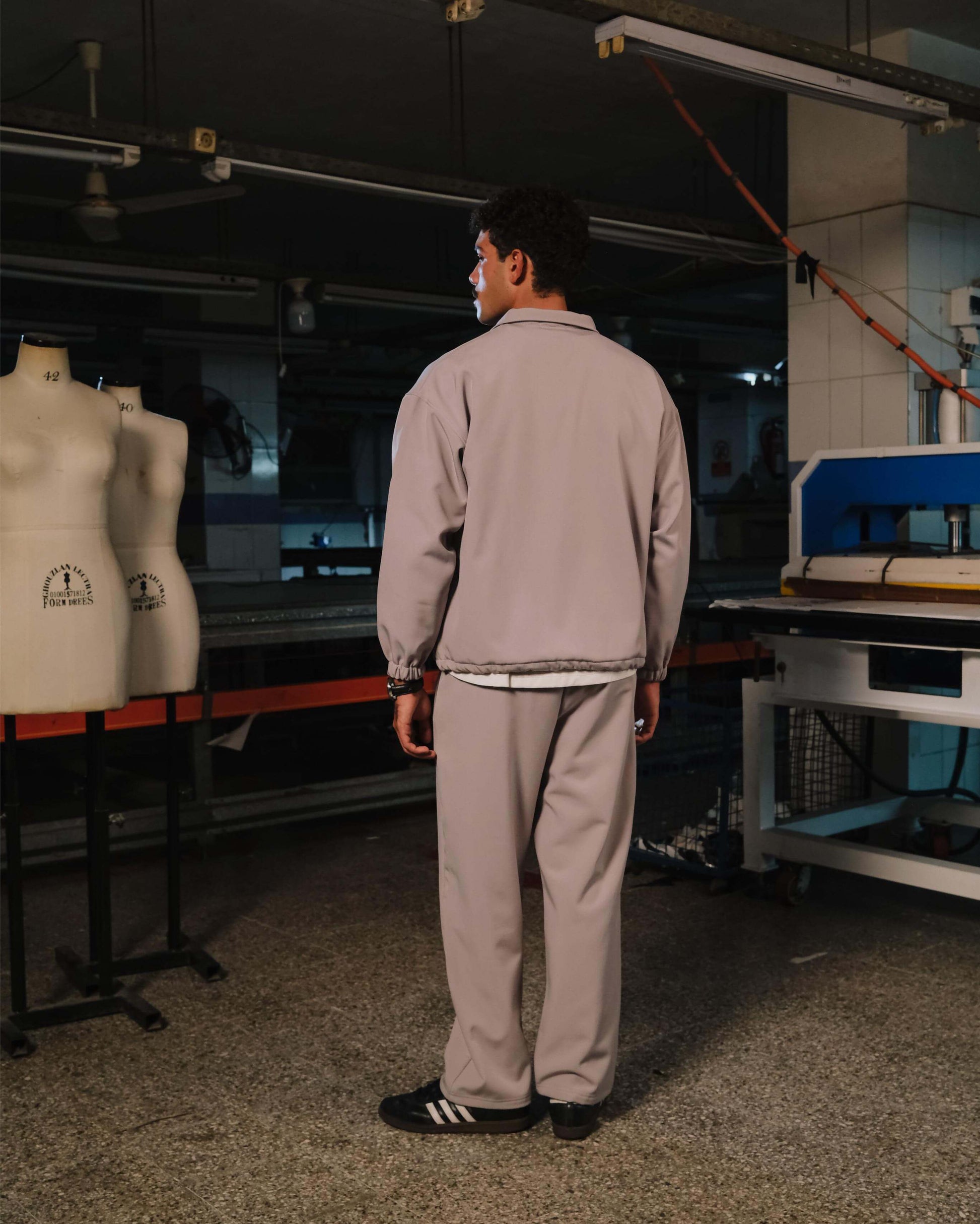 Person in a gray outfit standing in a workshop with mannequins and equipment.