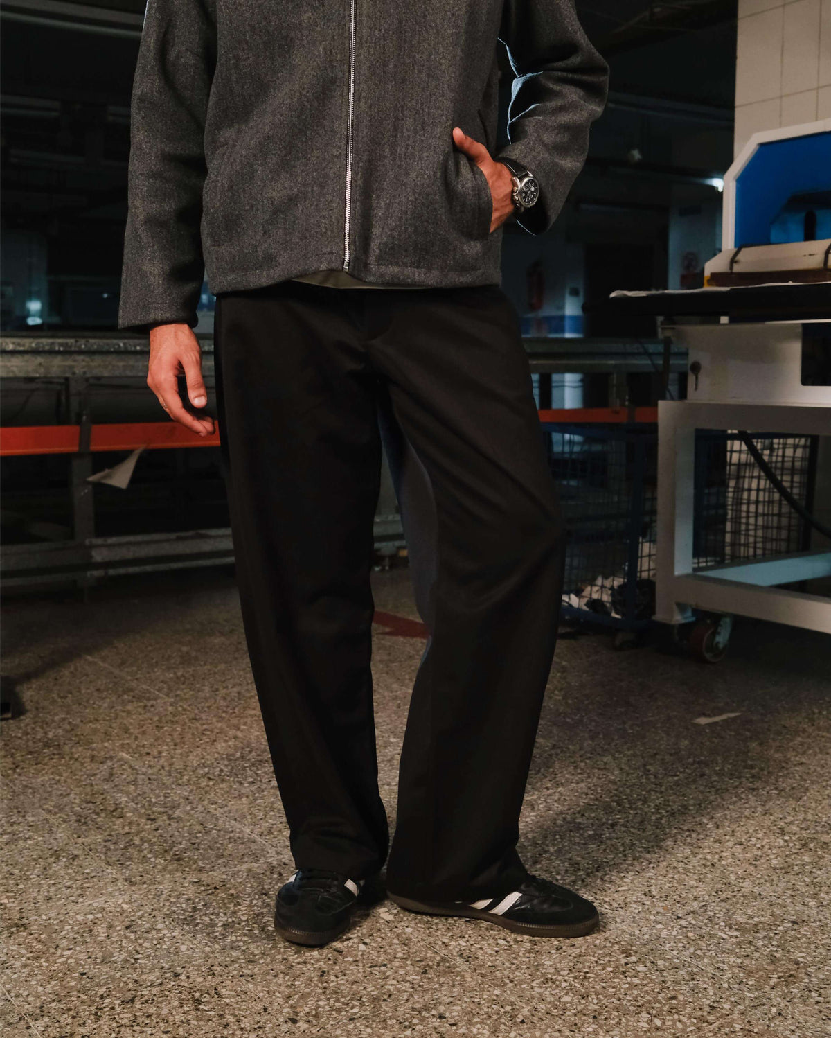 Person wearing a gray jacket and black pants standing in an industrial setting.