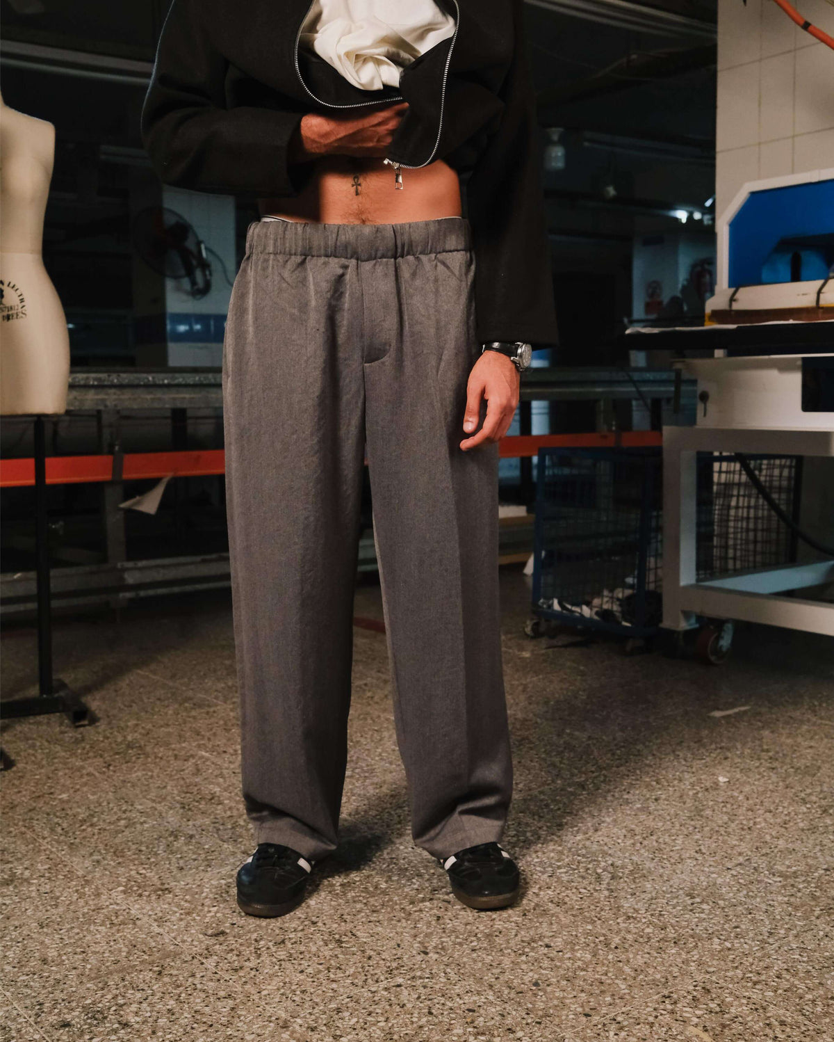 Person wearing gray pants and black shoes in an indoor setting with equipment and a mannequin in the background.
