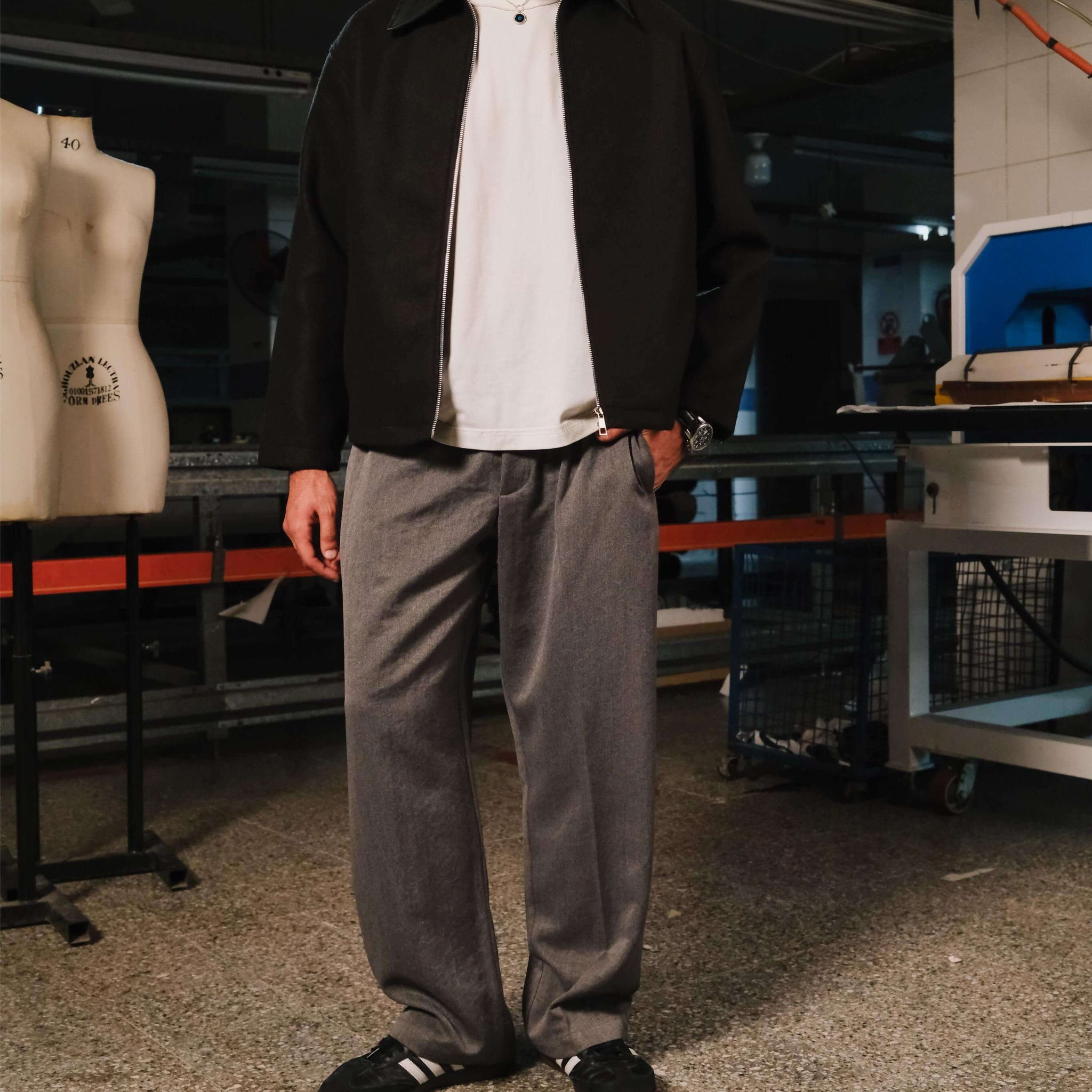 Man wearing a black jacket, white shirt, and gray pants standing in an indoor setting with equipment and mannequins in the background.