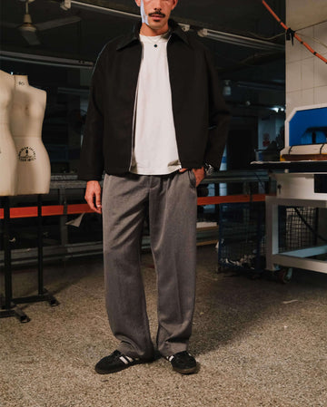 Man wearing a black jacket, white shirt, and gray pants standing in an indoor setting with equipment and mannequins in the background.