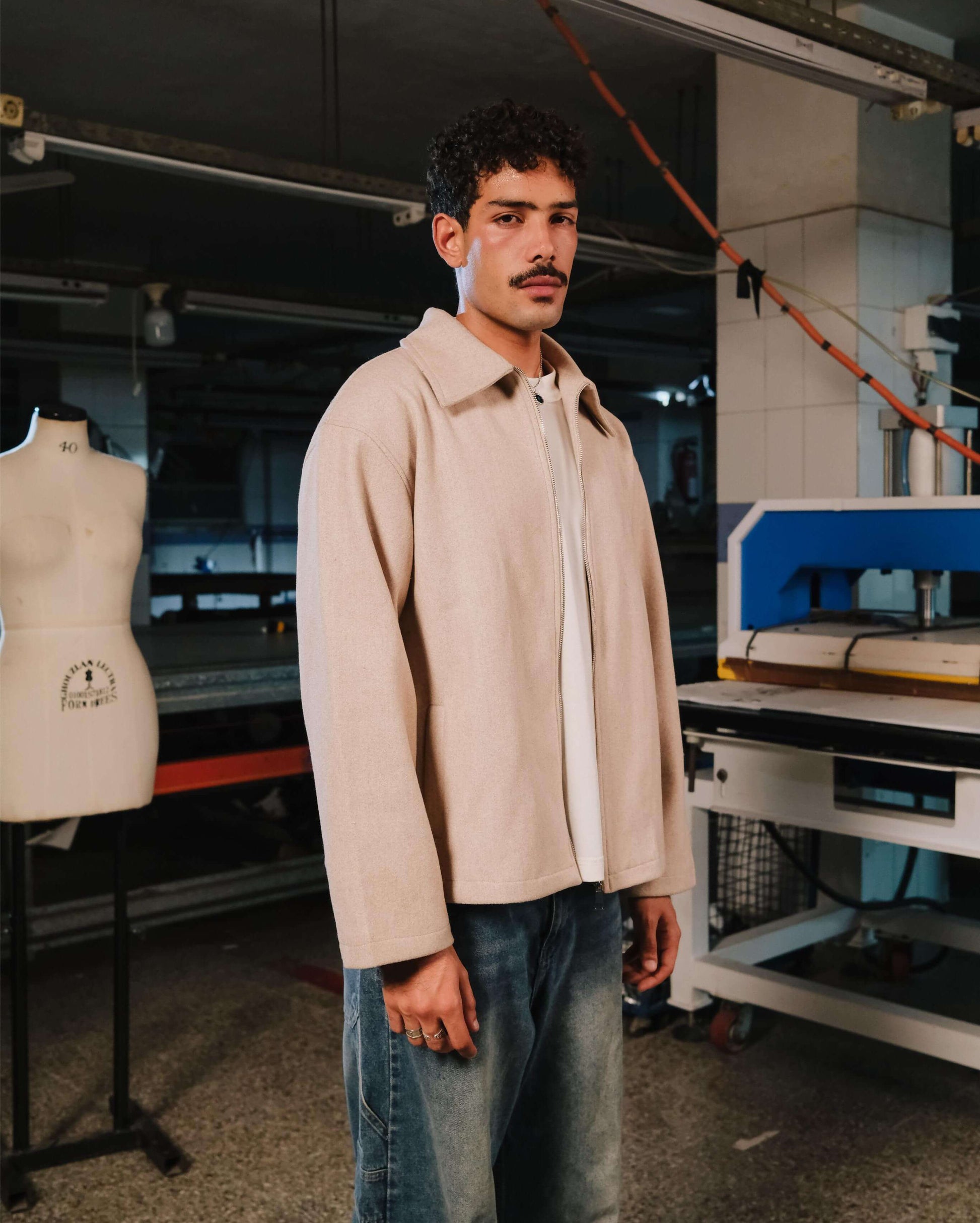 Man wearing a beige jacket in a workshop setting with sewing machines and mannequins.