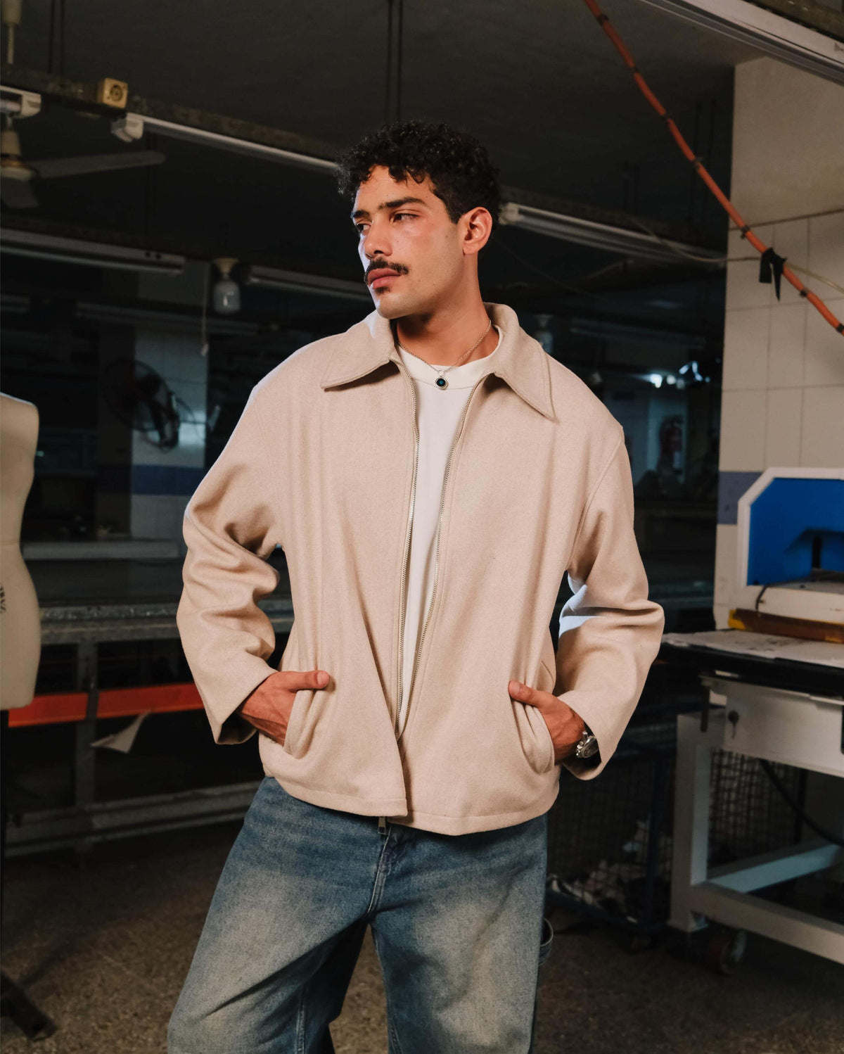 Man wearing a beige jacket and blue jeans standing in an industrial setting.