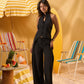 A woman modeling a black sleeveless halter top and matching wide-leg pants in a beach-themed setting with striped lounge chairs, a yellow-and-white umbrella, and warm sandy tones.