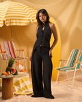 A woman modeling a black sleeveless halter top and matching wide-leg pants in a beach-themed setting with striped lounge chairs, a yellow-and-white umbrella, and warm sandy tones.
