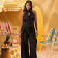 A model stands in a beach-themed studio setting wearing a black sleeveless halter top with a front tie and matching wide-leg pants. The scene includes a yellow-and-white striped umbrella, striped lounge chairs, a surfboard, and a small table with fruit and towels.