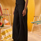 A model wearing a black sleeveless top and matching wide-leg trousers, standing in a beach-themed setting with yellow walls and striped lounge chairs.
