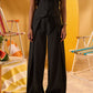 A model wearing a black sleeveless vest and matching wide-leg trousers with a drawstring waist, standing in a warm yellow studio setting with beach chairs and a towel in the background.