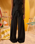 A model wearing a black sleeveless vest and matching wide-leg trousers with a drawstring waist, standing in a warm yellow studio setting with beach chairs and a towel in the background.