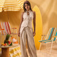 A woman stands in a beach-themed studio setting wearing a light beige sleeveless halter top with front ties and matching wide-leg pants. She is posed between striped beach chairs, with a yellow striped umbrella, surfboard, and fruit table in the background.