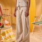 A model wearing a light beige sleeveless vest and matching wide-leg trousers with a drawstring waist, standing in a beach-themed setting with yellow walls and striped lounge chairs.