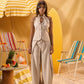 A woman stands in a beach-themed studio setting wearing a light beige sleeveless halter top with front buttons and matching wide-leg pants. She is posed between striped beach chairs and a yellow-and-white umbrella against a warm beige backdrop.
