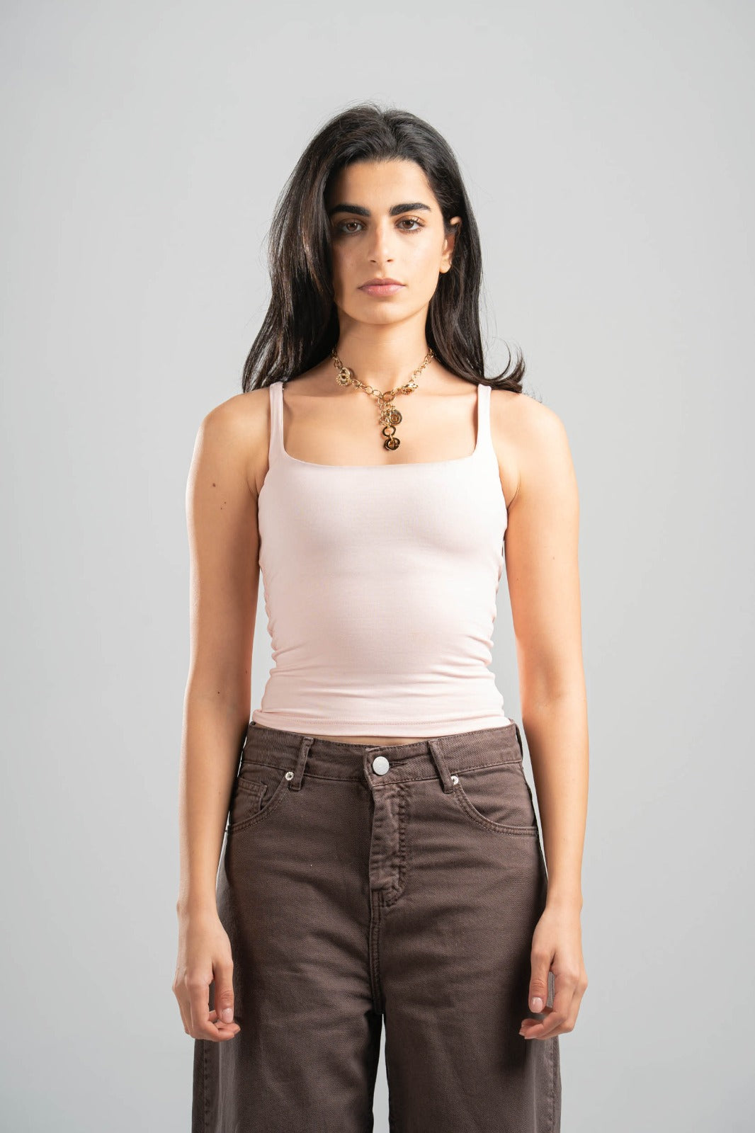 Woman wearing a light pink tank top and brown pants against a gray background