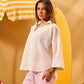 Woman in a white blouse and pink pants standing under a yellow and white striped umbrella with a yellow surfboard on a beige background.