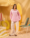 Woman in a pink shirt and white pants standing in a colorful beach-themed setting with chairs and an umbrella.