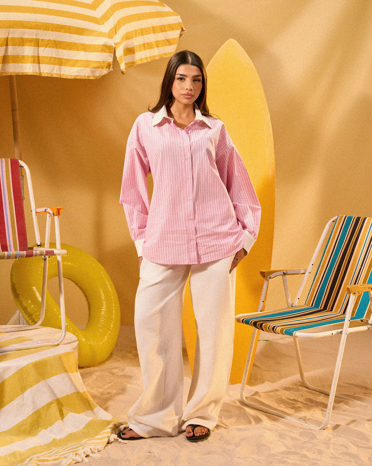 Woman in a pink shirt and white pants standing in a colorful beach-themed setting with chairs and an umbrella.