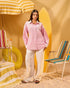 Woman in a pink shirt and white pants standing in a colorful beach-themed setting with chairs and an umbrella.