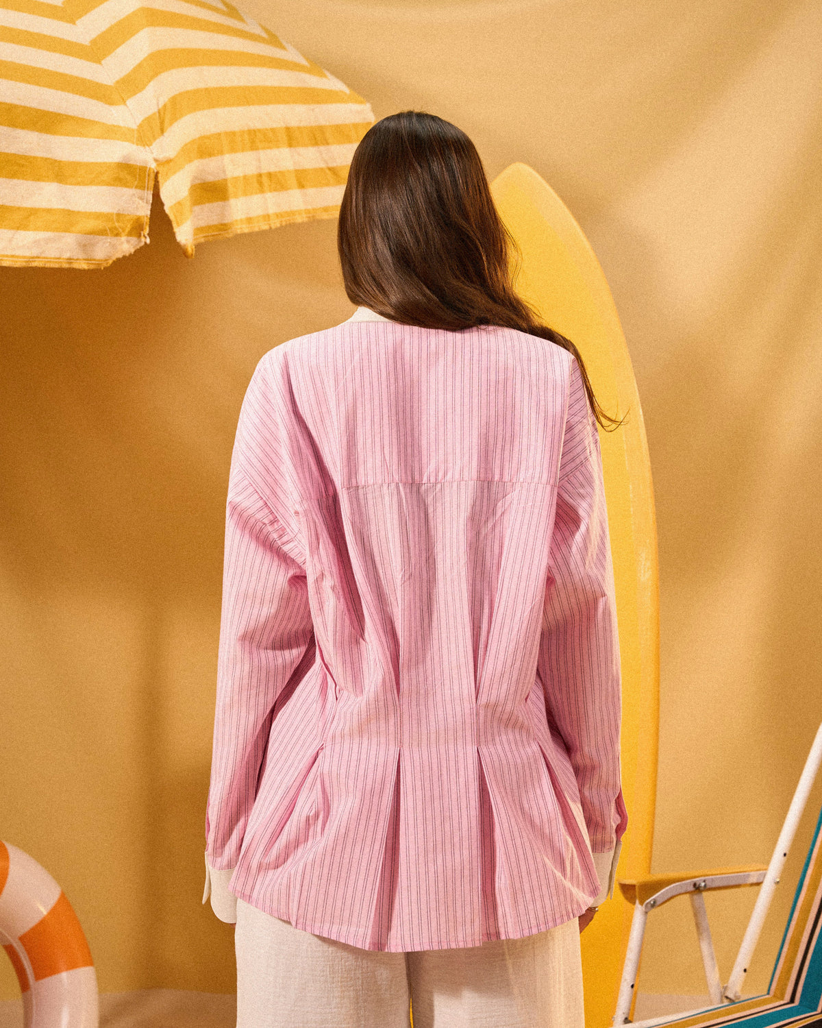 Person wearing a pink striped shirt against a yellow background with a surfboard and umbrella.