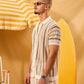 Man in a patterned shirt standing next to a yellow and white striped umbrella and surfboard on a yellow background
