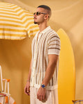 Man in a patterned shirt standing next to a yellow and white striped umbrella and surfboard on a yellow background