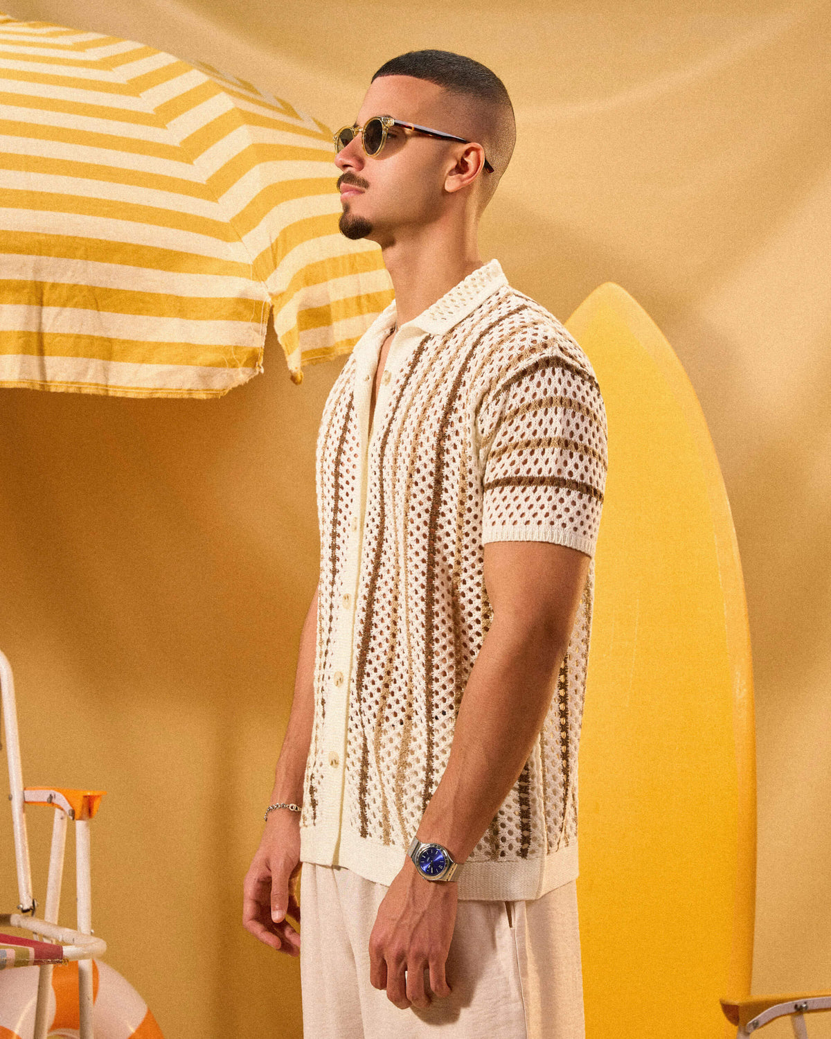 Man in a patterned shirt standing next to a yellow and white striped umbrella and surfboard on a yellow background