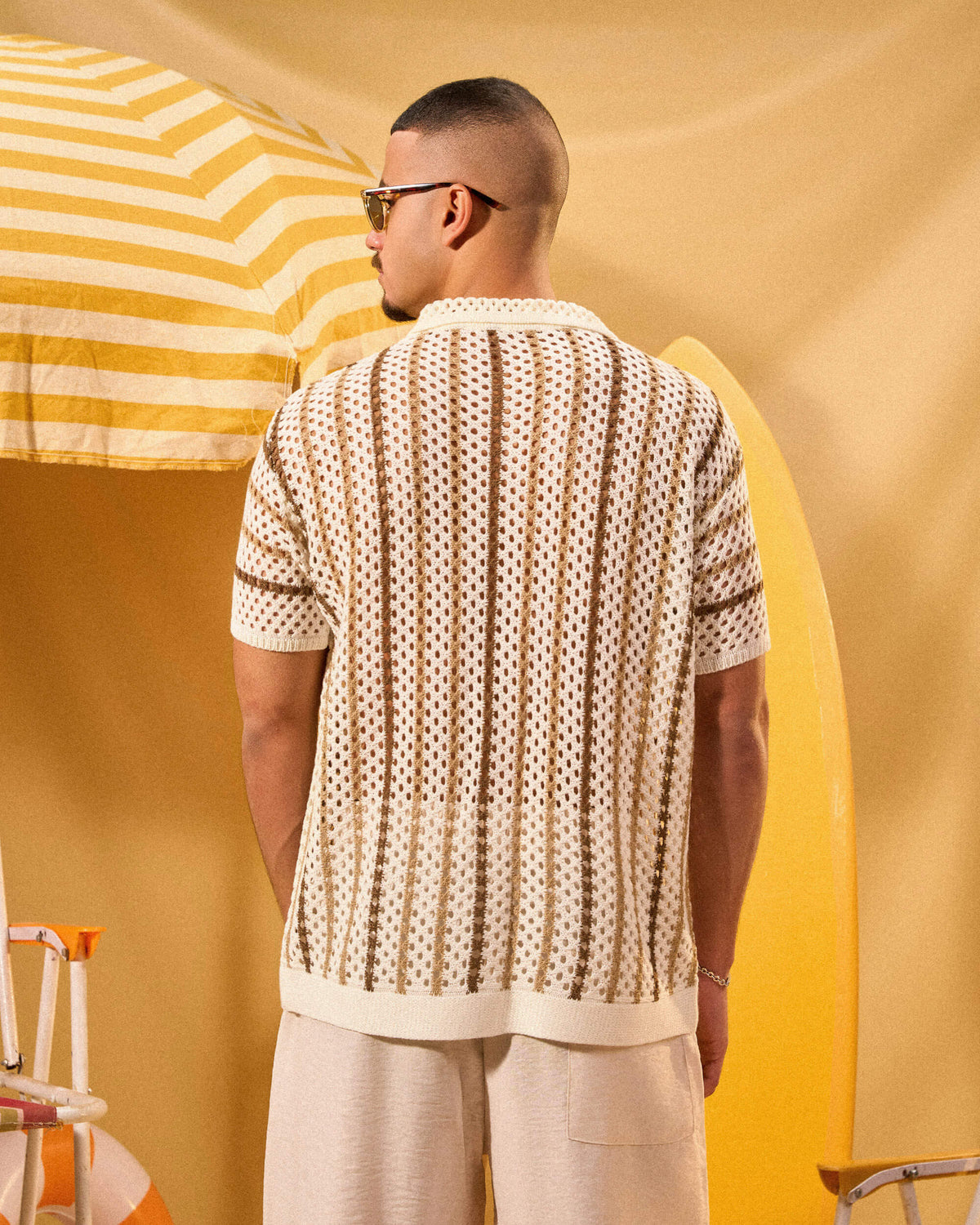 Man wearing a patterned shirt and sunglasses standing under a yellow and white striped umbrella.